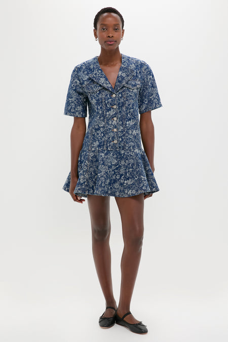 A person stands against a white background wearing a GANNI short-sleeve blue floral fit-and-flare dress and black ballet flats.