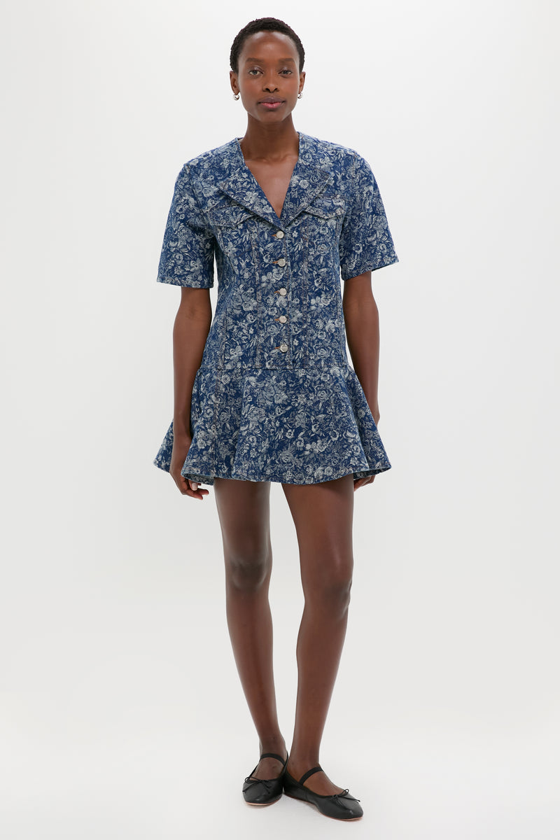 A person stands against a white background wearing a GANNI short-sleeve blue floral fit-and-flare dress and black ballet flats.