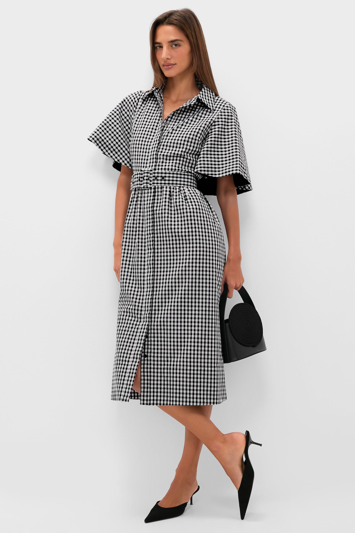 A woman wears a Tuckernuck black and white gingham midi shirt dress with cape sleeves, paired with a small black handbag and black pointed-toe heels.