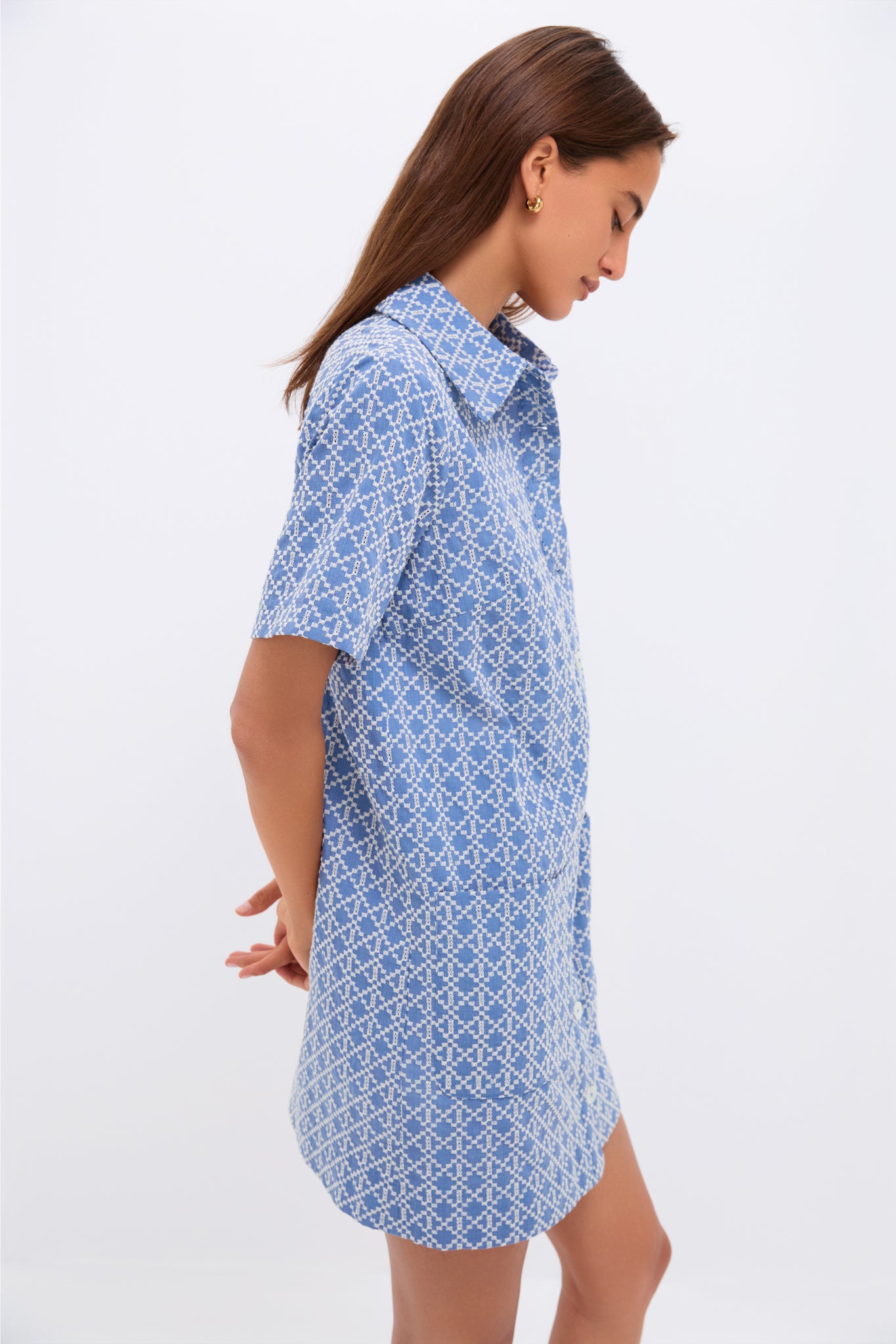 A woman models a blue and white patterned vintage-inspired mini dress by Pomander Place, standing sideways with her hands behind her back against a plain white background.
