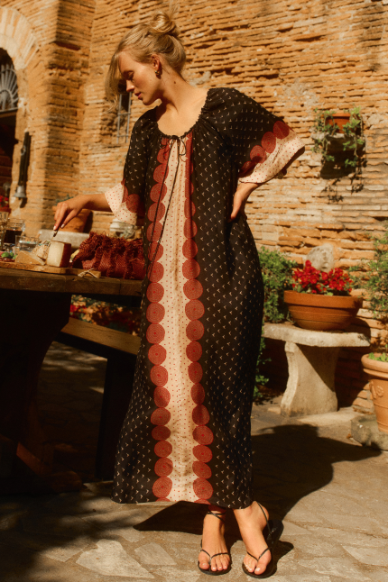 Woman leaning over a table in front of a brick wall in a black and rust printed maxi dress. 