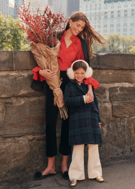 A woman stands on a city park bridge with a little girl. The woman wears a black blazer, a red blouse, black pants, and black flats while carrying a set of paper wrapped florals. The little girl wears a tartan patterned coat, cream pants, gold t-strap shoes, and ear muffs.