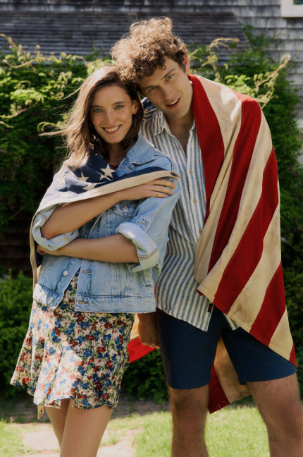 Man and woman standing together with an American flag wrapped around them. Woman is in a jean jacket and floral dress and man in a striped shirt and navy shorts.