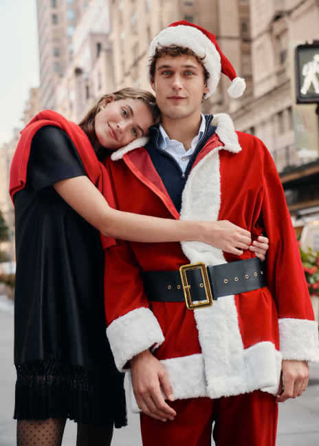 On the left, a woman stands wearing a black dupioni mini dress with tassels, a red sweater wrapped around her shoulders, and polka dot tights. On the right, a man wears a Santa Claus costume over top a navy quarter zip and blue button down.