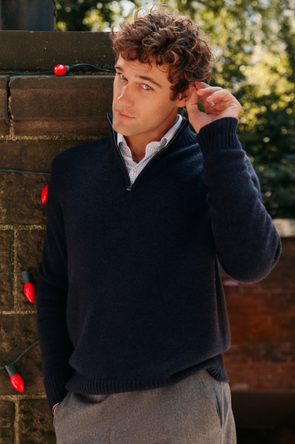 A man leans against a brick pillar, wearing a navy quarter zip and gray trousers.