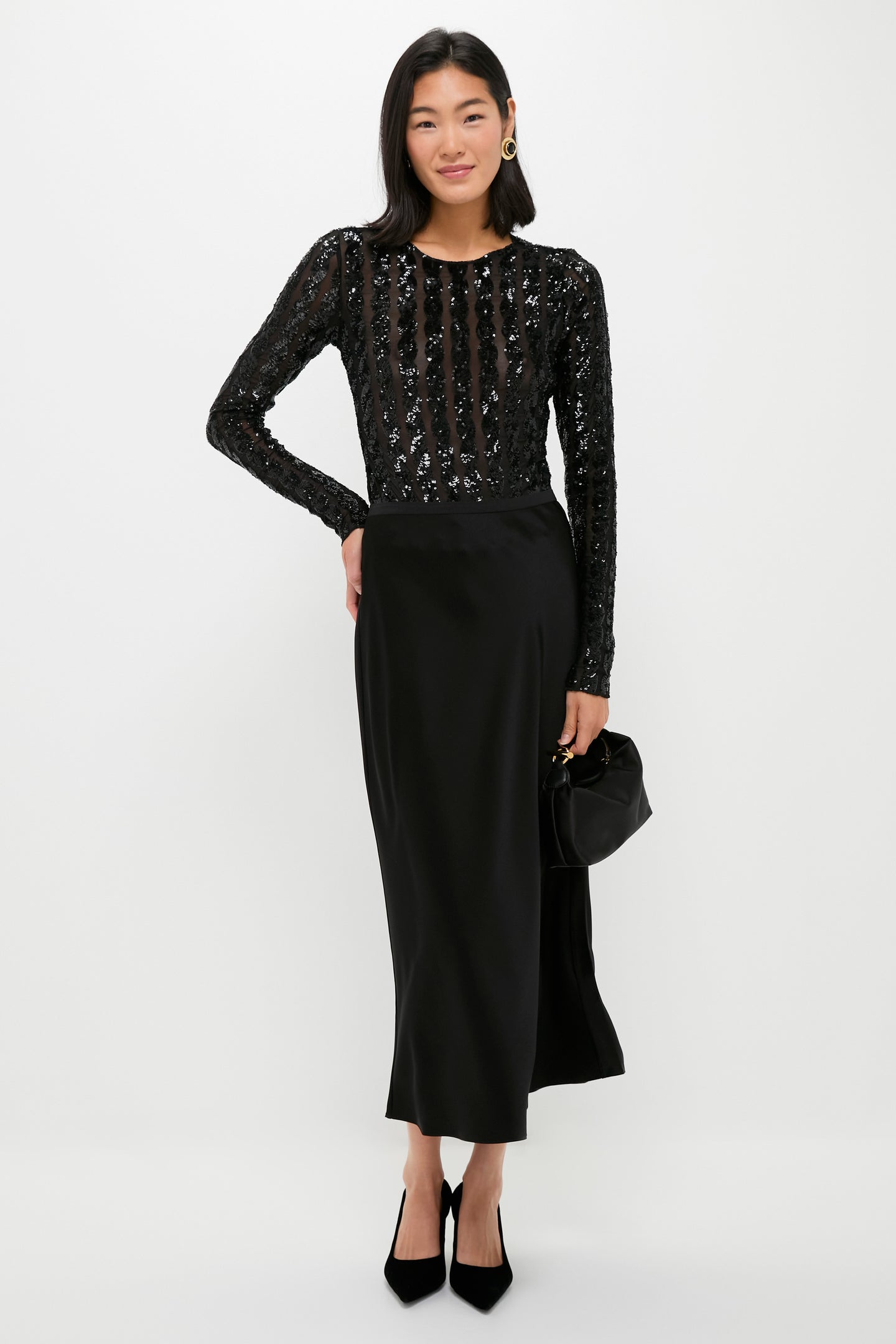 A woman stands against a white background wearing a long-sleeved black sequin top, long black skirt, black heels, and holding a sleek Liffner calf leather clutch—an elegant, Scandinavian-inspired evening look.