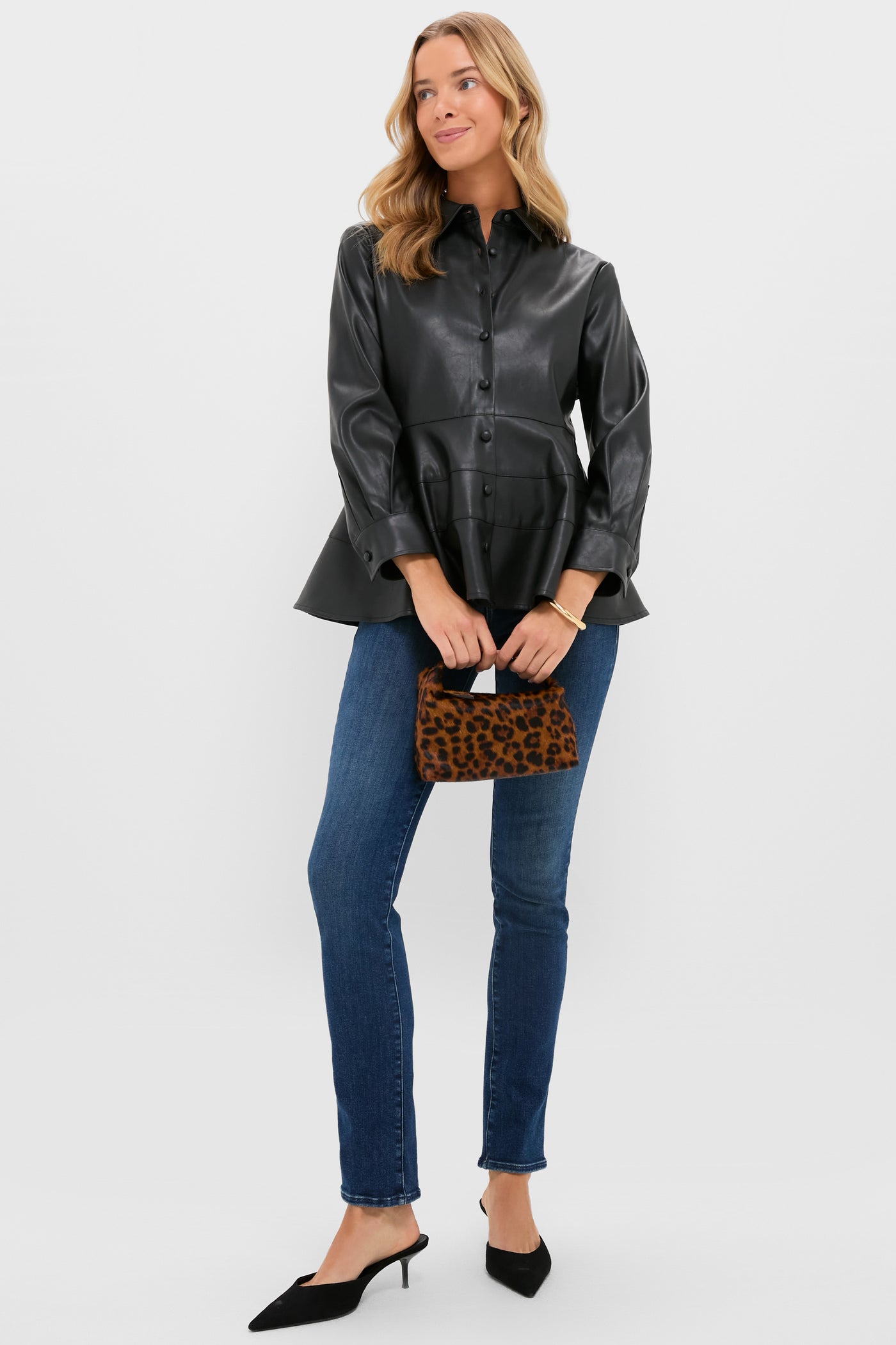 A woman in a Pomander Place black leather button-down blouse, blue jeans, black pointed heels, and holding a small leopard print handbag stands against a plain light background.
