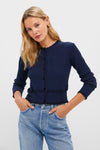 A woman wears a navy blue Tuckernuck Lily cardigan with scallop ruffle trim, velvet buttons, and black accents, paired with light blue jeans against a plain light background.