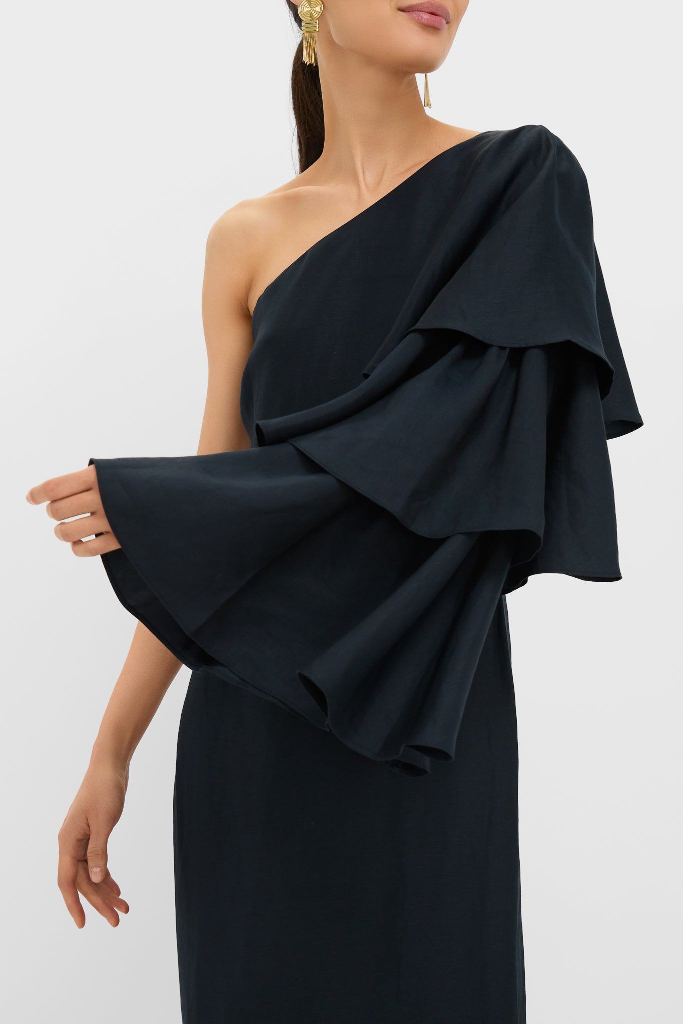 Woman in Black Bidi Bidi Bom Bom Tea Length Dress with asymmetric neckline, dramatic ruffled sleeve, accessorized with gold drop earrings; a classic, effortless, high-end look.