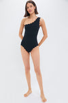 A woman stands barefoot in a scalloped-edge black one-shoulder swimsuit by Marysia, embodying high-fashion swimwear style.