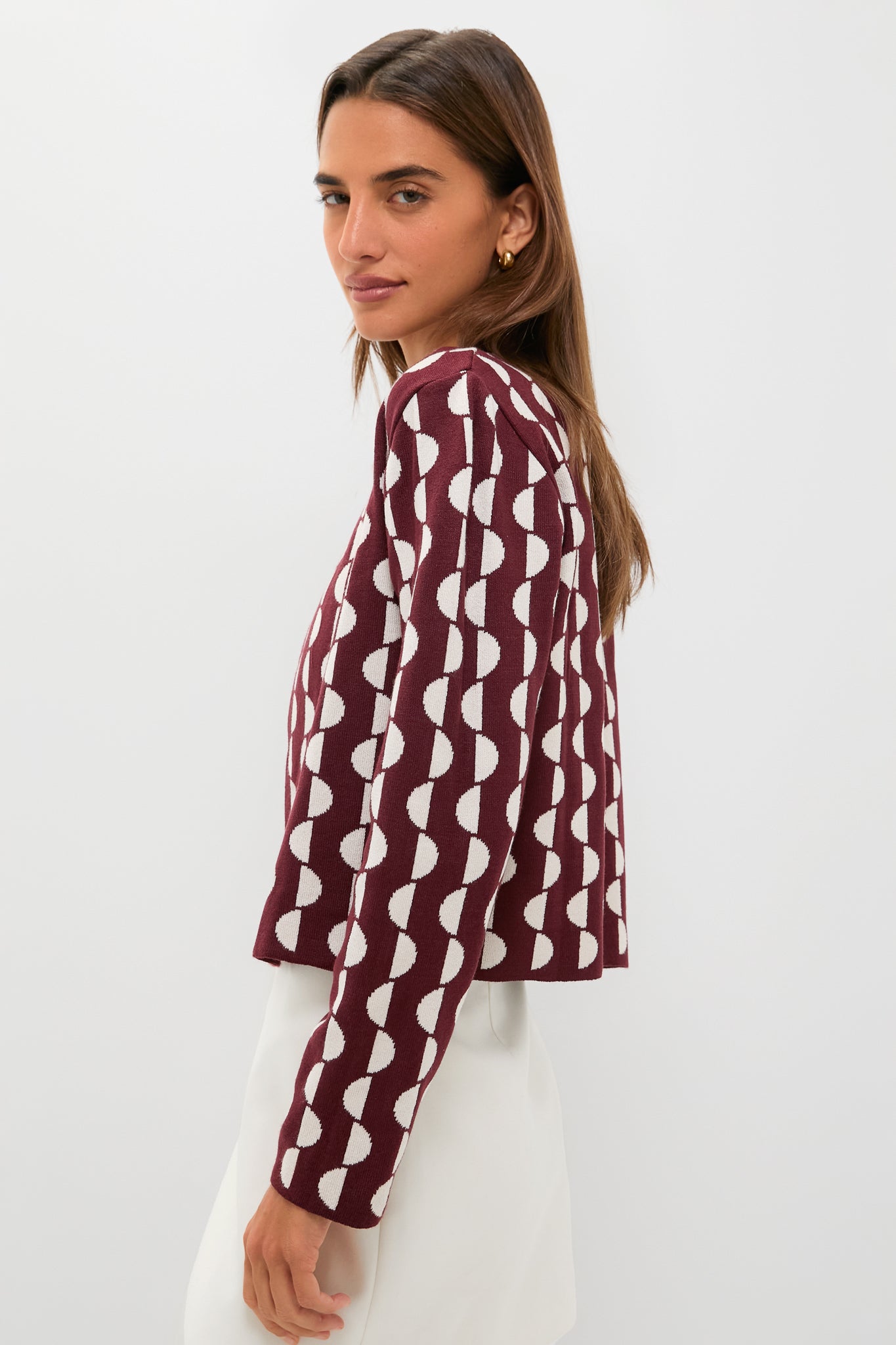 A woman stands against a plain light background, wearing a maroon long-sleeve knit jacket with a white geometric pattern and subtle hook-and-eye closure by Dress To, paired with a white skirt.