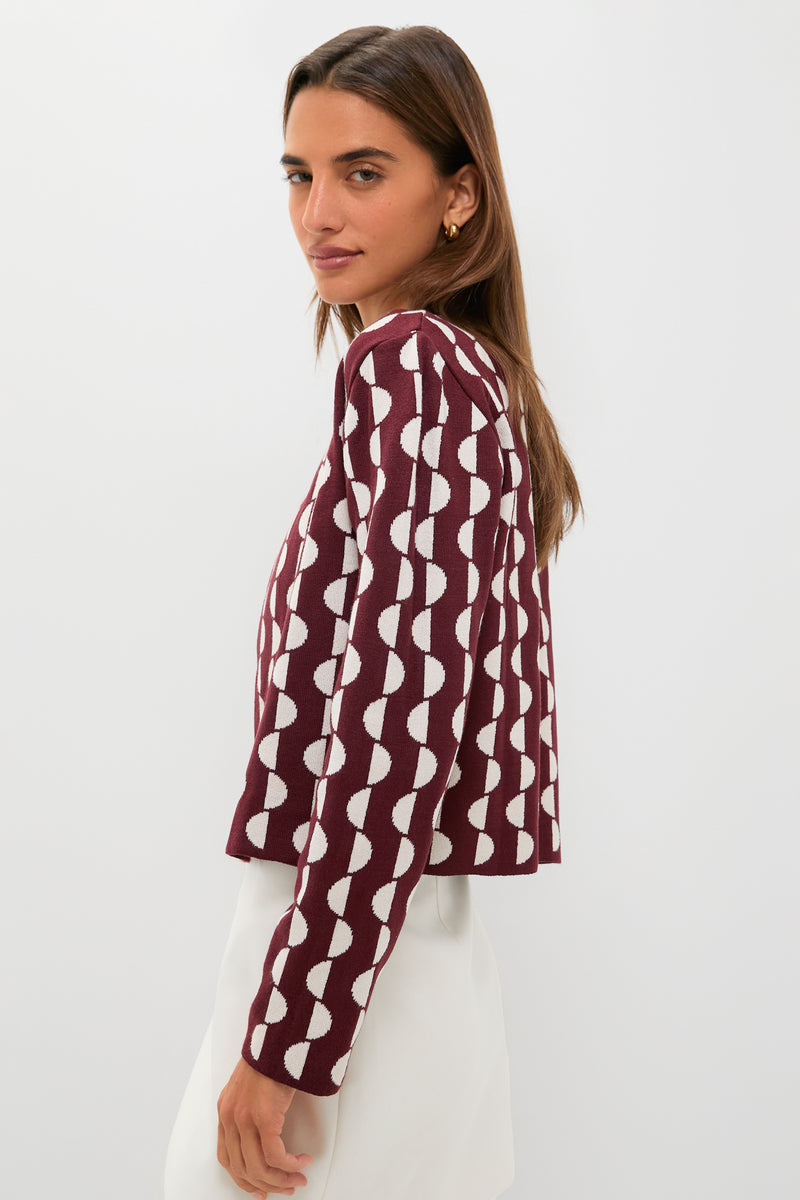 A woman stands against a plain light background, wearing a maroon long-sleeve knit jacket with a white geometric pattern and subtle hook-and-eye closure by Dress To, paired with a white skirt.