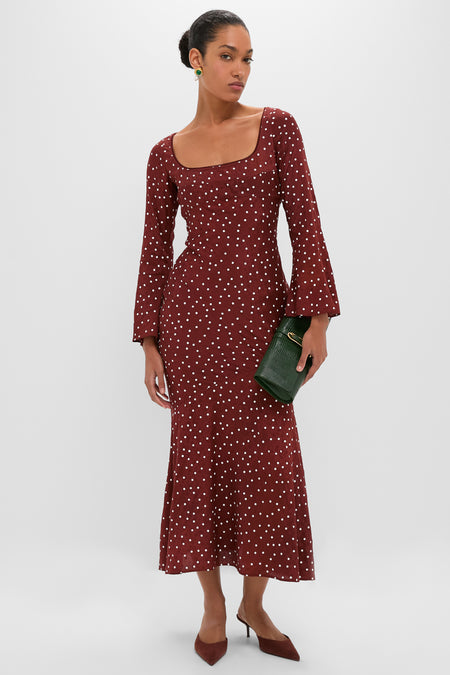 Woman in a burgundy Burnt Umber Narah Maxi Dress with polka dots, holding a dark-green clutch, embodying classic, effortless style with fun, high-end details like flared sleeves.