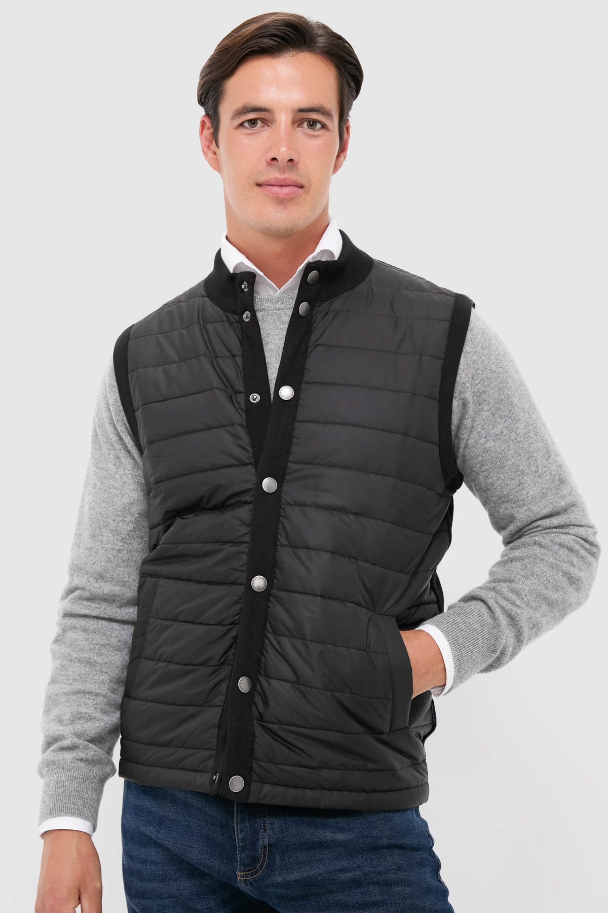 A man in a gray sweater, blue jeans, and a black Barbour Essential Gilet stands confidently in men's outerwear.