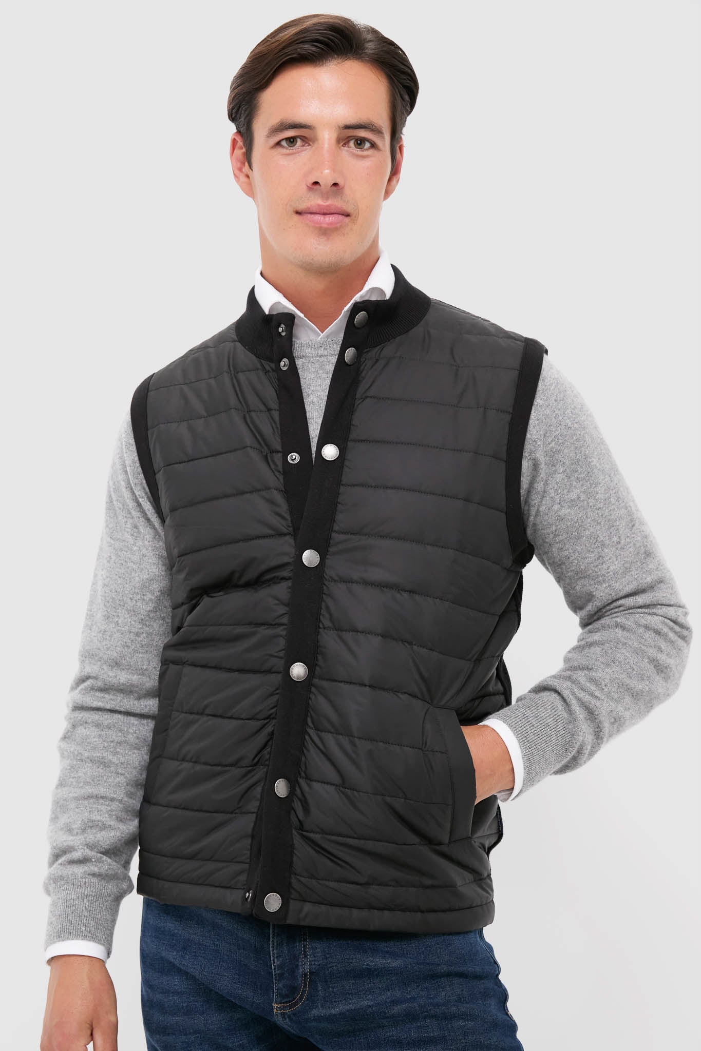 A man in a gray sweater, blue jeans, and a black Barbour Essential Gilet stands confidently in men's outerwear.