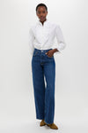 A woman stands against a white background wearing a white Polo Ralph Lauren cotton-silk shirt with ruffled details, blue wide-leg jeans, and brown shoes.