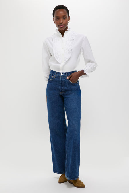 A woman stands against a white background wearing a white Polo Ralph Lauren cotton-silk shirt with ruffled details, blue wide-leg jeans, and brown shoes.