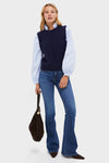 A woman in a navy sweater vest layered over a light blue ruffle-collar blouse with puffed sleeves, flared jeans, heeled boots, and carrying a large black bag.