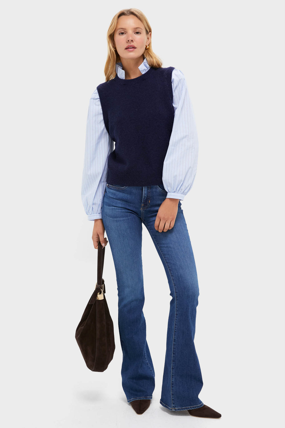 A woman in a navy sweater vest layered over a light blue ruffle-collar blouse with puffed sleeves, flared jeans, heeled boots, and carrying a large black bag.
