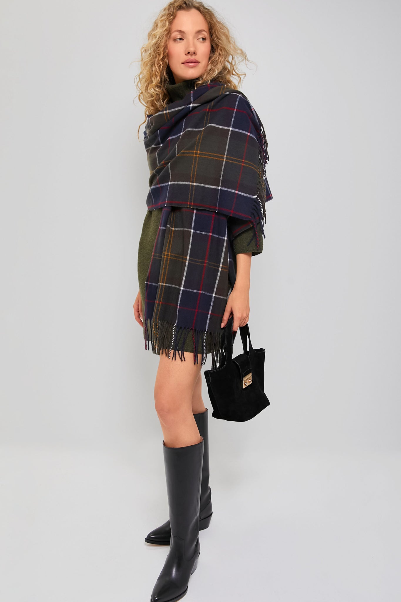 A woman wearing a Barbour tartan wrap over a green dress, black knee-high boots, and holding a small black handbag stands against a plain light gray background.