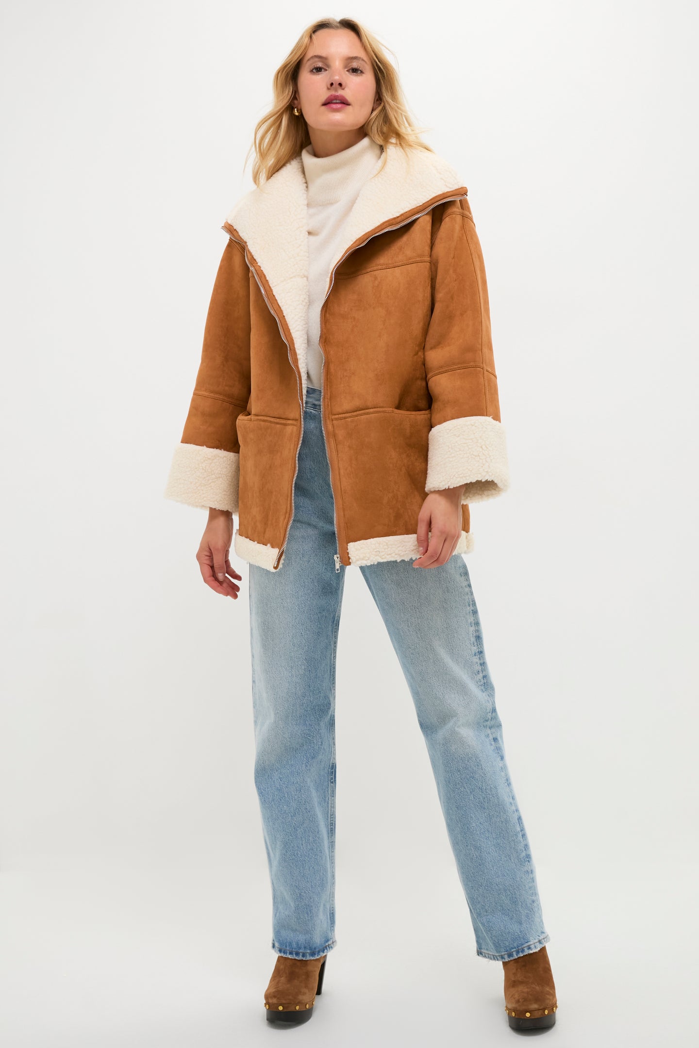 A woman stands facing forward wearing a Moon River camel suede coat with faux fur trim, a light turtleneck sweater, light blue jeans, and brown heeled shoes against a plain white background.