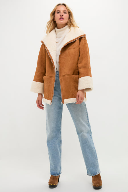 A woman stands facing forward wearing a Moon River camel suede coat with faux fur trim, a light turtleneck sweater, light blue jeans, and brown heeled shoes against a plain white background.