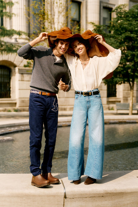 Man and woman standing on the edge of a fountain with a suede jacket over their heads. Man in a gray sweater and jeans. Woman is in a white blouse and jeans.
