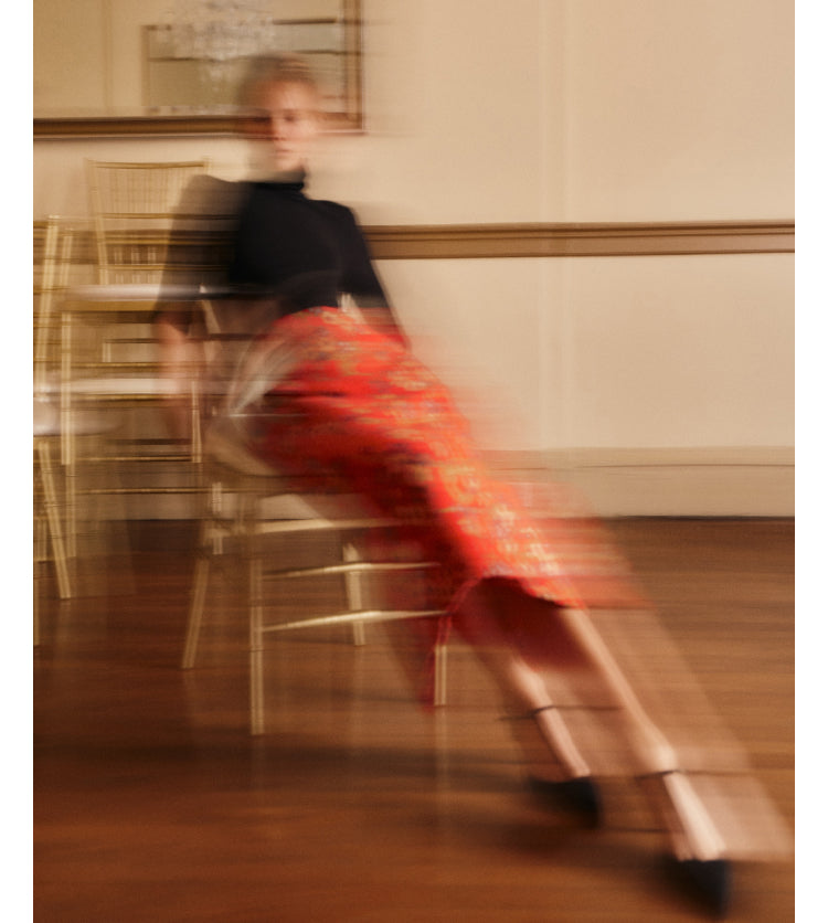 A woman wearing a red floral jacquard skirt, black top, and black t-strap heels sitting on a chair.