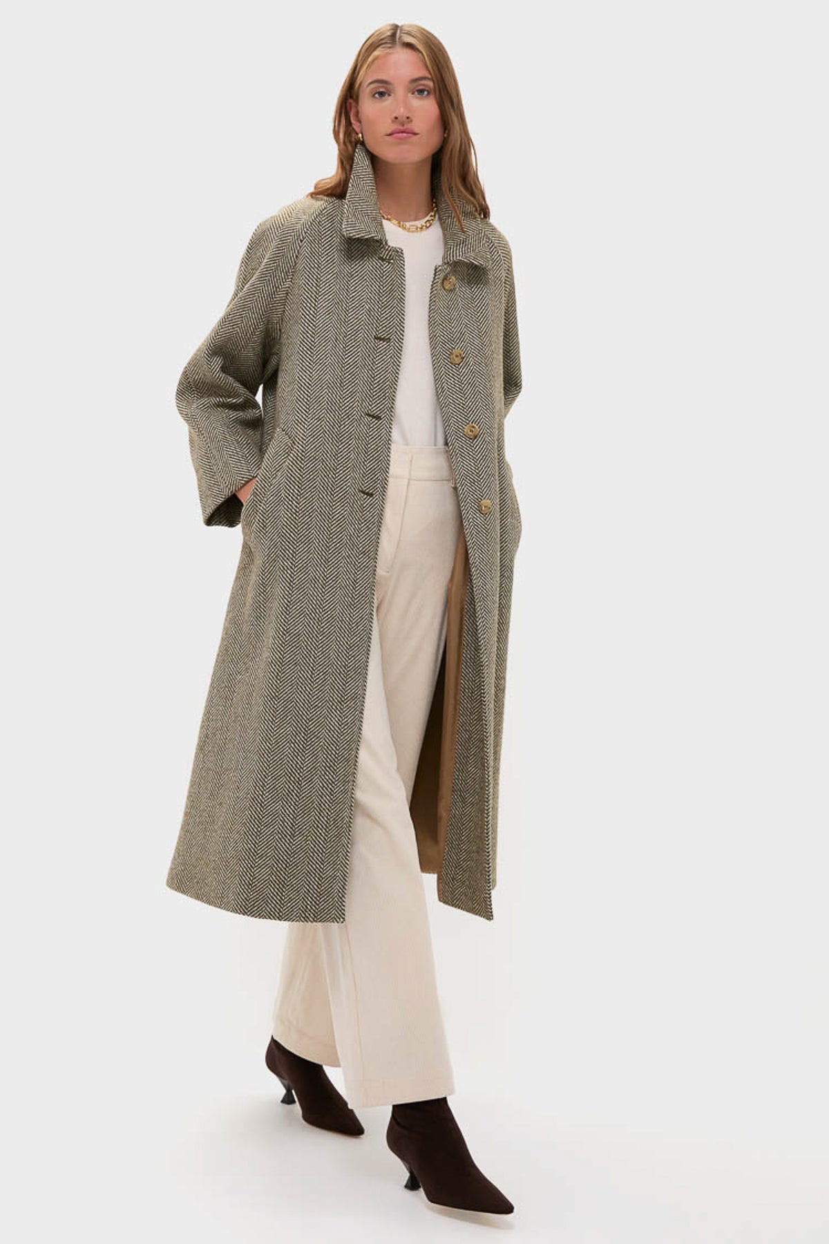 A woman wearing a Weekend Max Mara oversized wool coat with a classic herringbone pattern over a white top and cream trousers, styled with black ankle boots and hands in her coat pockets.