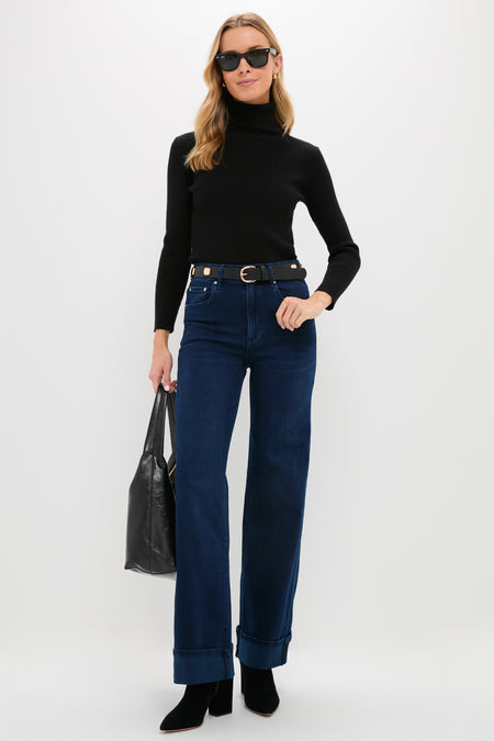 A woman wearing a black turtleneck, LE JEAN Los Angeles-made dark indigo high-rise jeans, a black belt and boots, sunglasses, and holding a black tote bag stands against a plain white background.