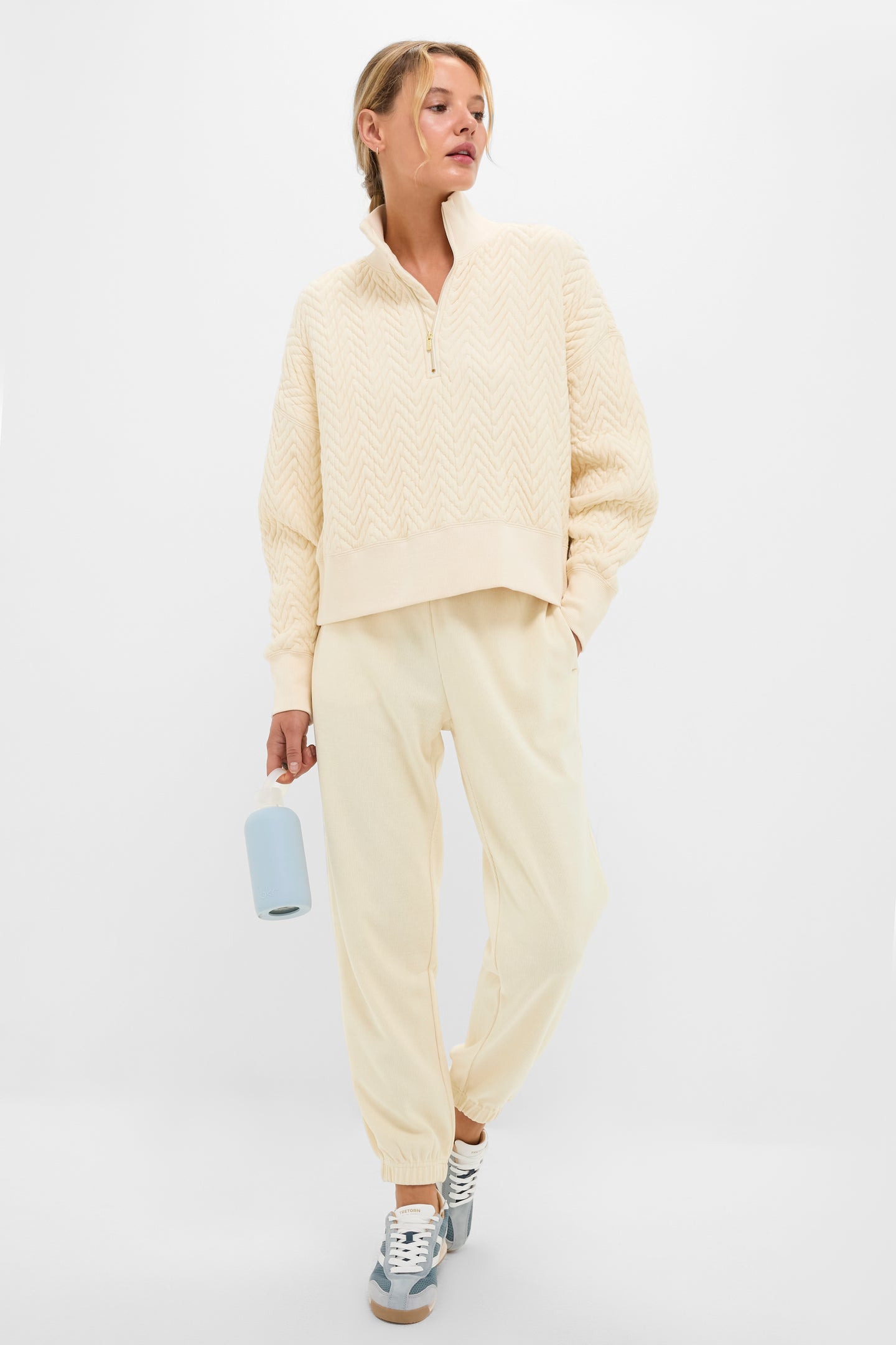 A person dressed in Tnuck Sport's cream-colored quilted pullover with a mock collar and matching jogger pants, paired with grey sneakers and holding a water bottle, stands against a plain light background.