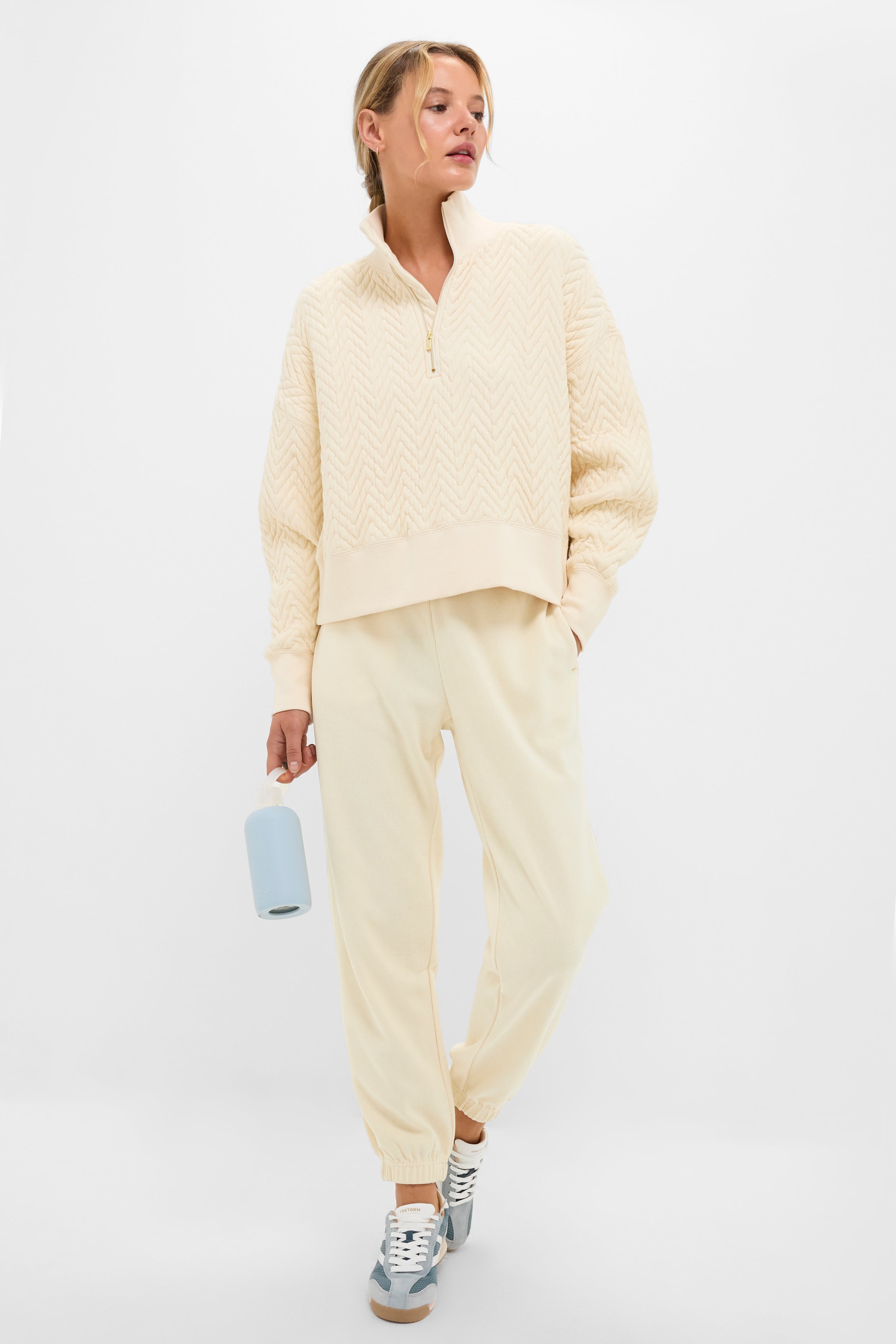 A person dressed in Tnuck Sport's cream-colored quilted pullover with a mock collar and matching jogger pants, paired with grey sneakers and holding a water bottle, stands against a plain light background.