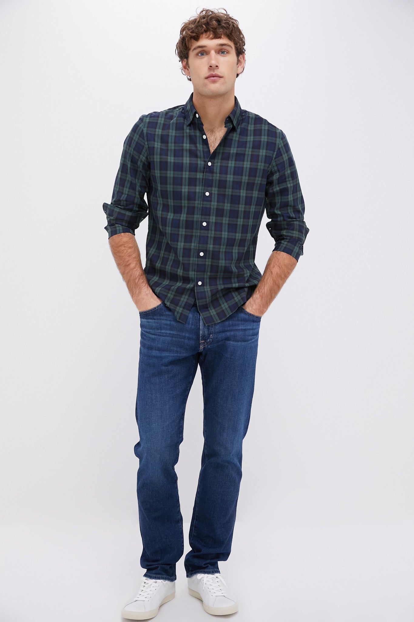 A man stands with hands in his pockets, wearing a Faherty dark green and navy plaid shirt made from an ultra-soft cotton blend, blue jeans, and white sneakers against a plain white background.