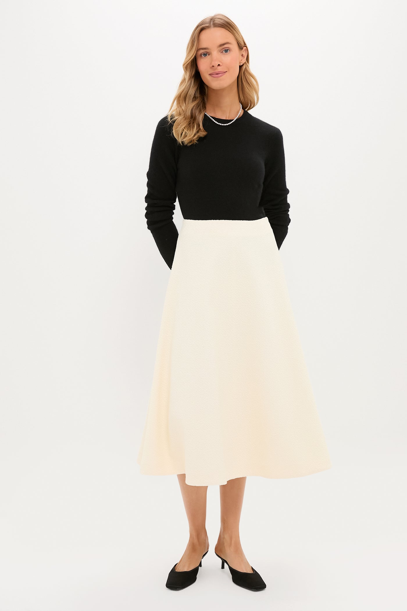 A woman stands against a plain background wearing a Tuckernuck black long-sleeve top, an A-line cream boucle skirt, black heels, and a necklace.