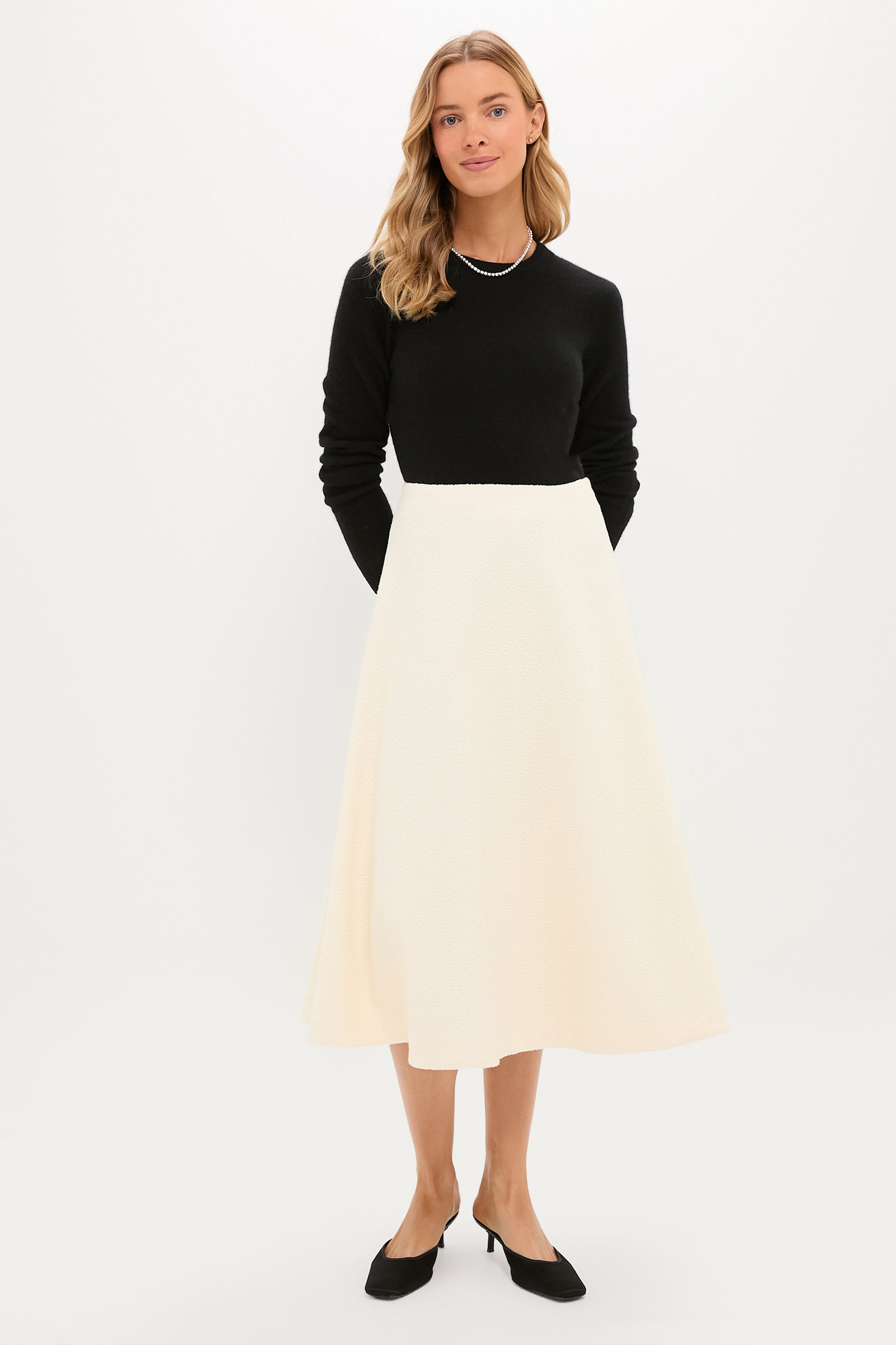 A woman stands against a plain background wearing a Tuckernuck black long-sleeve top, an A-line cream boucle skirt, black heels, and a necklace.