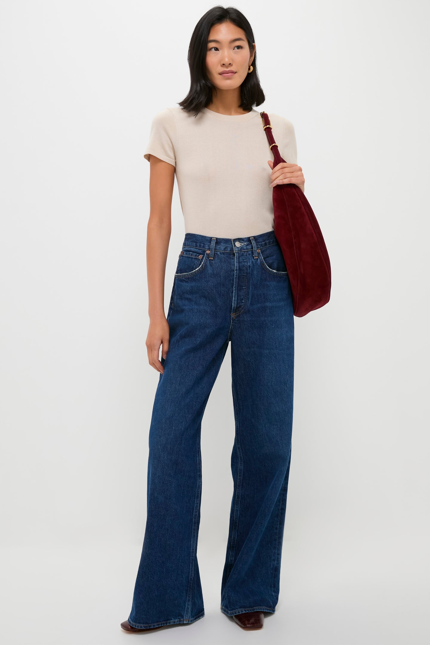 A woman stands against a plain background wearing a Tuckernuck beige relaxed fit crewneck t-shirt, wide-leg blue jeans, dark boots, and carrying a large dark red shoulder bag.