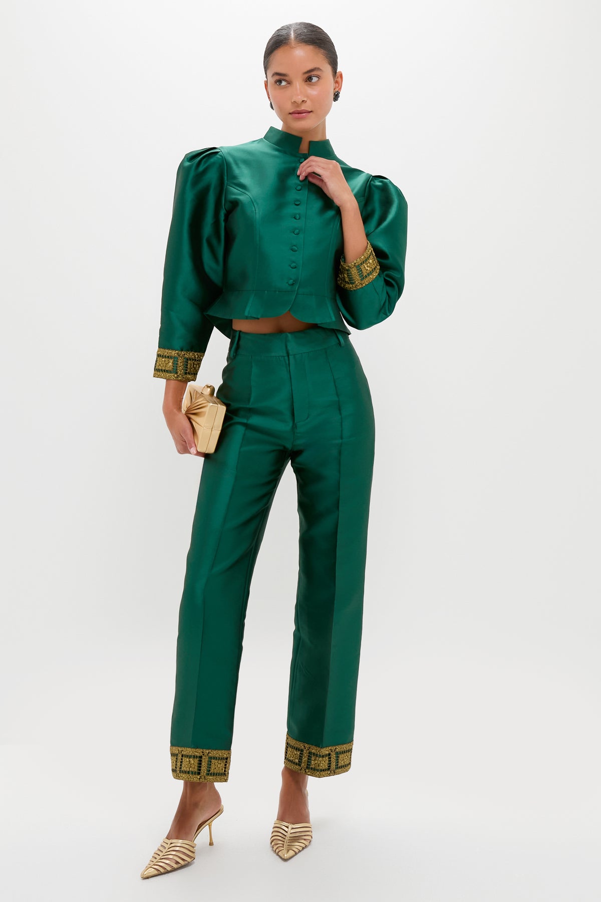 A woman wears a shiny green vintage-inspired DESTREE jacket and matching high-waisted pants with gold trim, holding a small beige clutch and wearing striped heels.