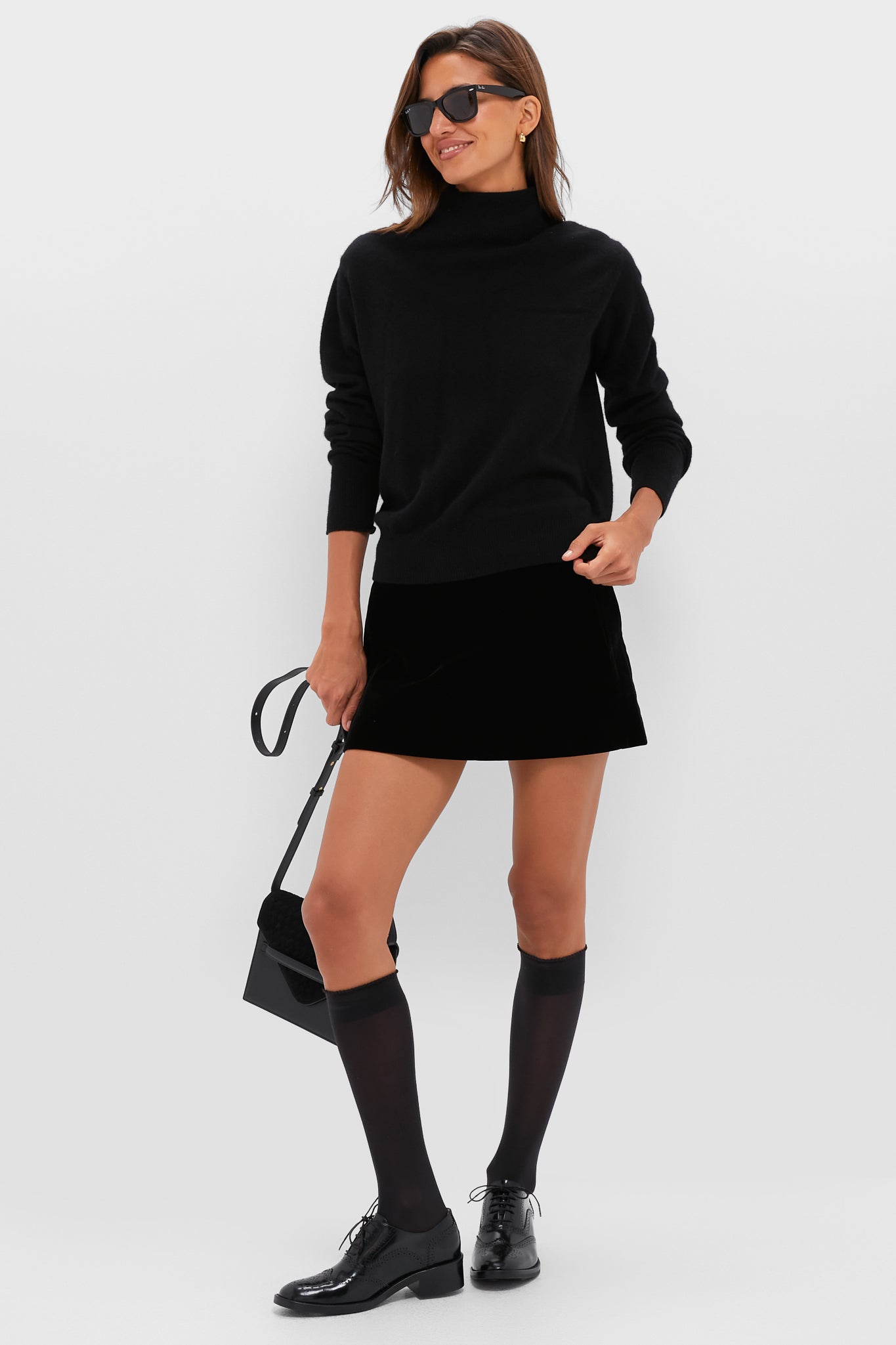 A woman wears a black turtleneck sweater, black mini skirt, knee-high socks, sunglasses, a small black handbag, and black leather loafers.