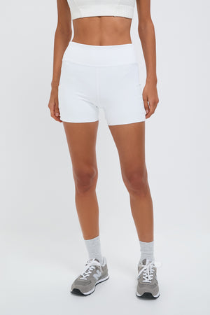 A person stands confidently wearing a white Tnuck Sport sports bra, white Tnuck Sport Aaron Tennis Short 4 Inch, gray socks, and gray sneakers.