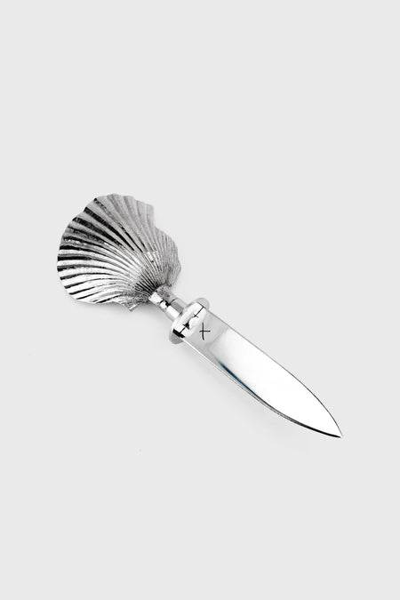 A Shelton Metal hand-carved oyster knife with a stainless steel blade and a scallop shell-shaped handle is displayed on a plain surface.