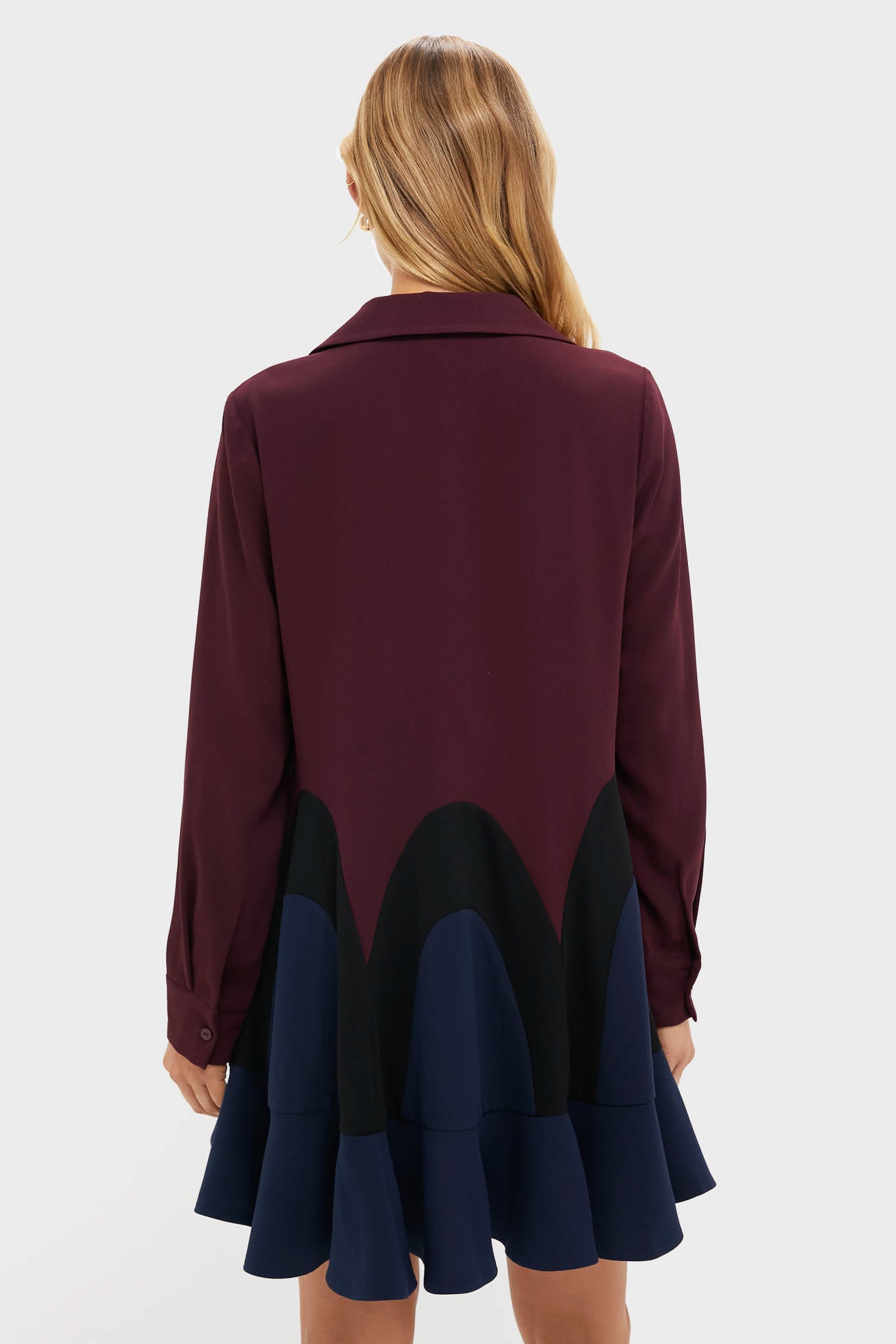 A woman faces away from the camera in a Tuckernuck long-sleeve burgundy shirt featuring black and navy scalloped patterns, a ruffled navy hem, and the playful swing silhouette of a flouncy mini dress.