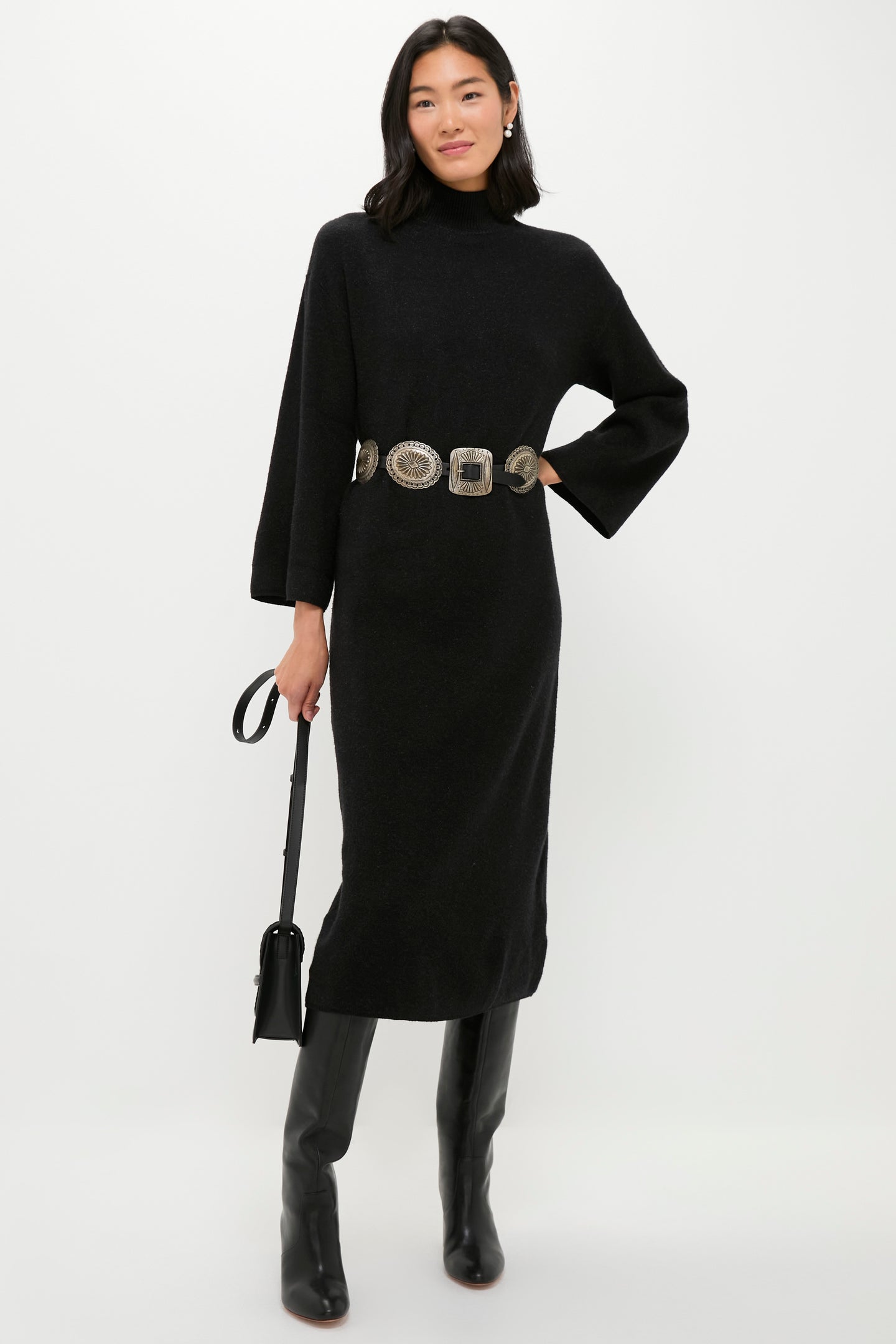 A woman in a long-sleeved black dress with a high neckline, wide belt, knee-high black boots, and black handbag, styled with elegant eliou freshwater pearl earrings, stands against a plain background.