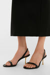 A person wearing Cult Gaia black heeled sandals with thin straps and gold stiletto heels stands on a white surface in a long black skirt.