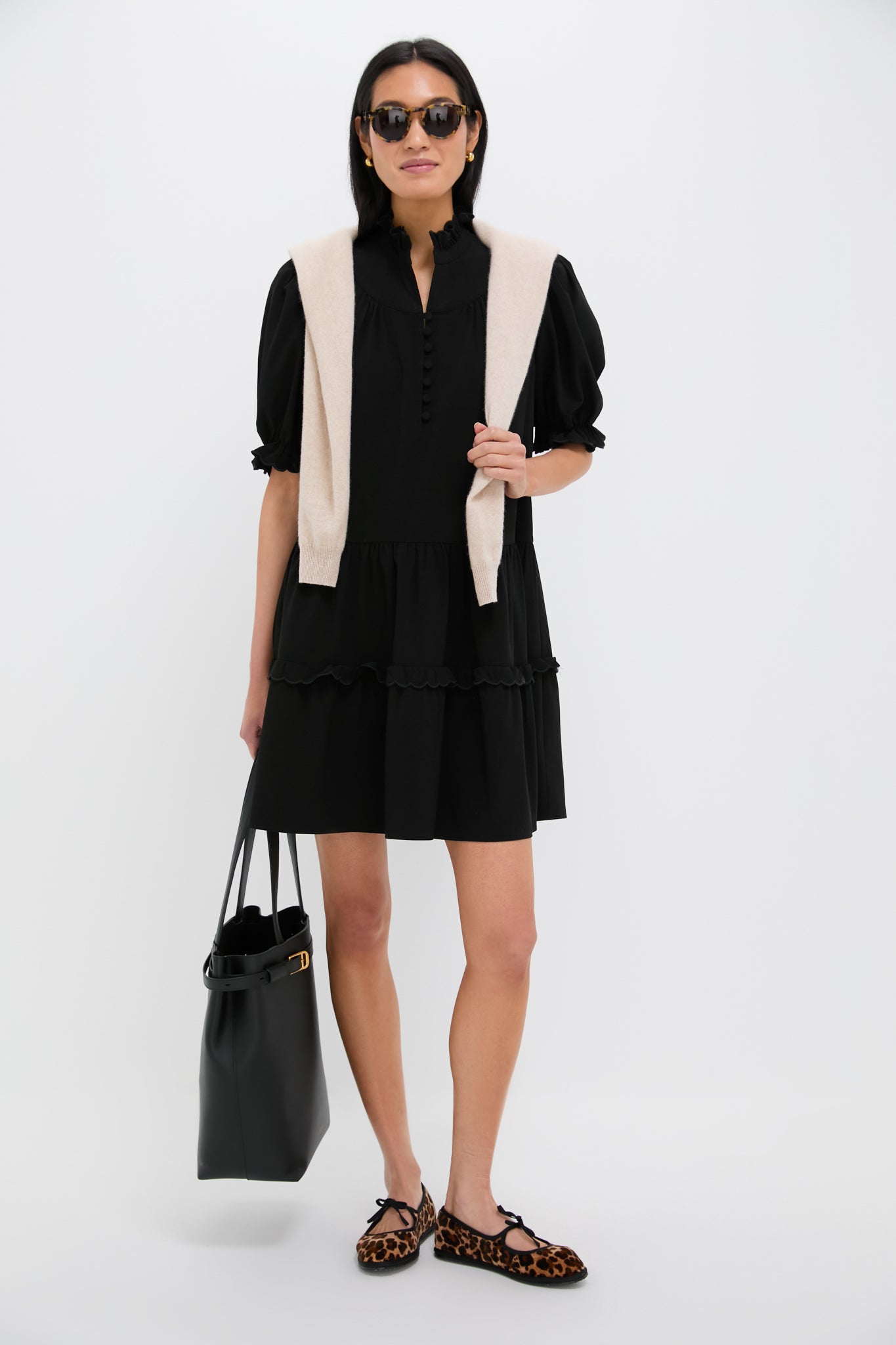 A woman stands against a white background wearing Pomander Places black puff sleeve mini dress, a beige sweater over her shoulders, sunglasses, leopard print flats, and holding a black tote bag.