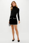 Young woman models the Black Velvet Rhinestone Loverly Mini Skirt with rhinestone trim, paired with a black turtleneck and slingback heels, exuding effortless high-end style.