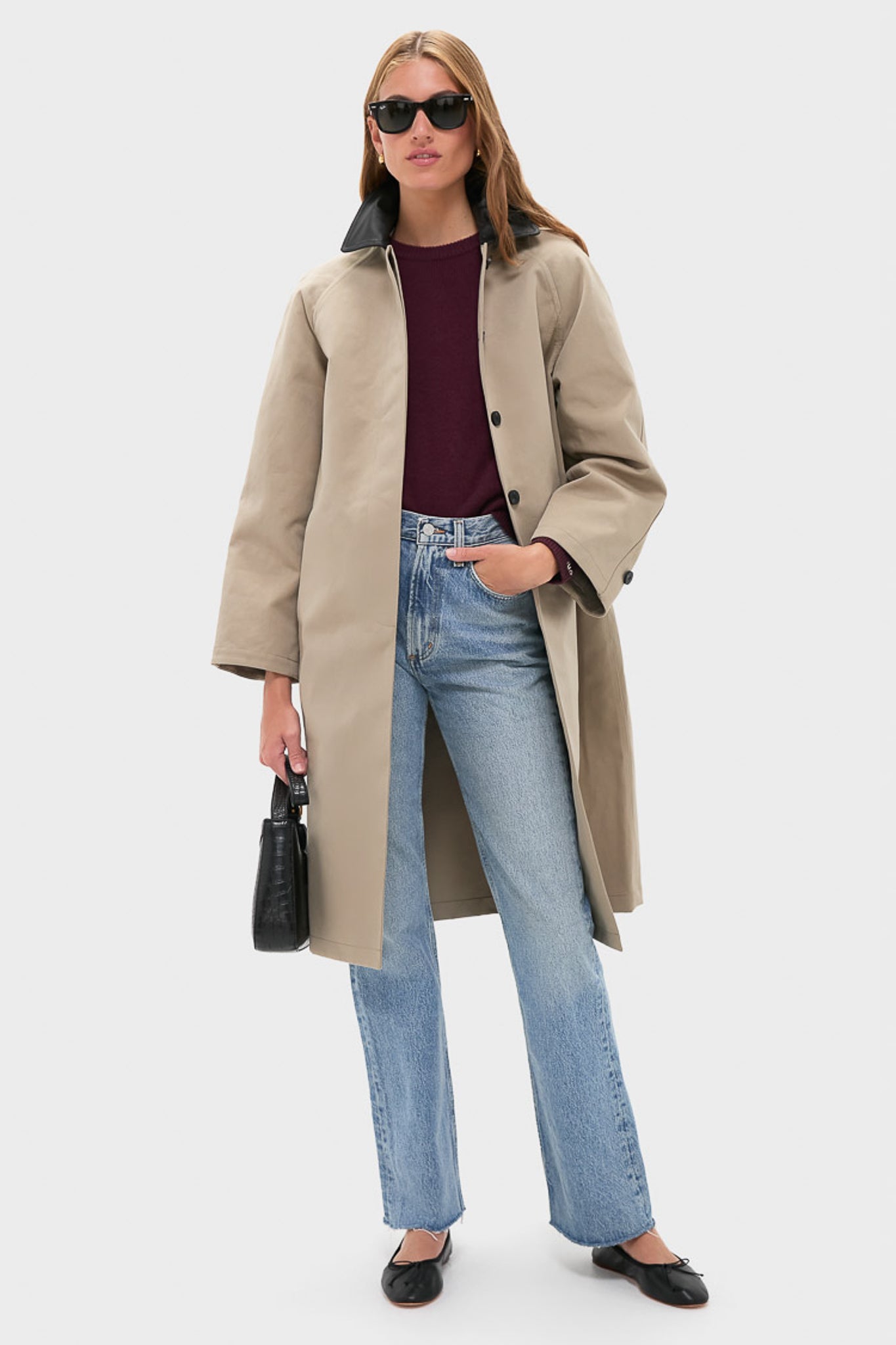 A woman in a STAUD Jonah Long Coat layers it over a burgundy top and blue jeans, paired with black flats, sunglasses, and a black handbag, standing against a plain background.