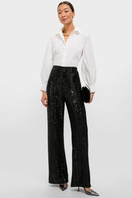 Woman in Black Sequin Tatum Pants paired with a classic white shirt, exuding effortless high-end style. She's accessorized with gold jewelry and a chic clutch.