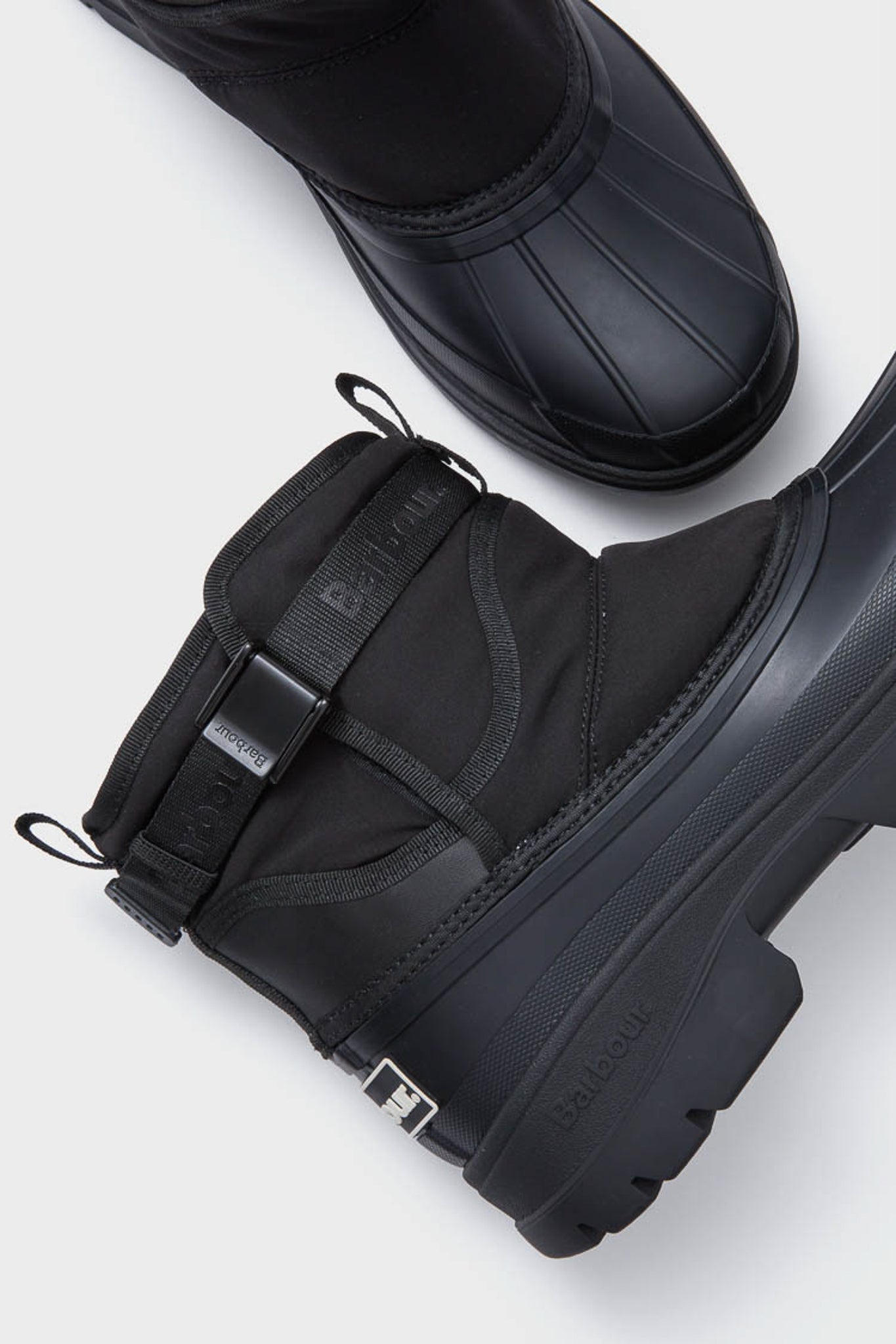 Two black Barbour waterproof winter boots with thick rubber soles and strap detail are displayed on a light gray background.