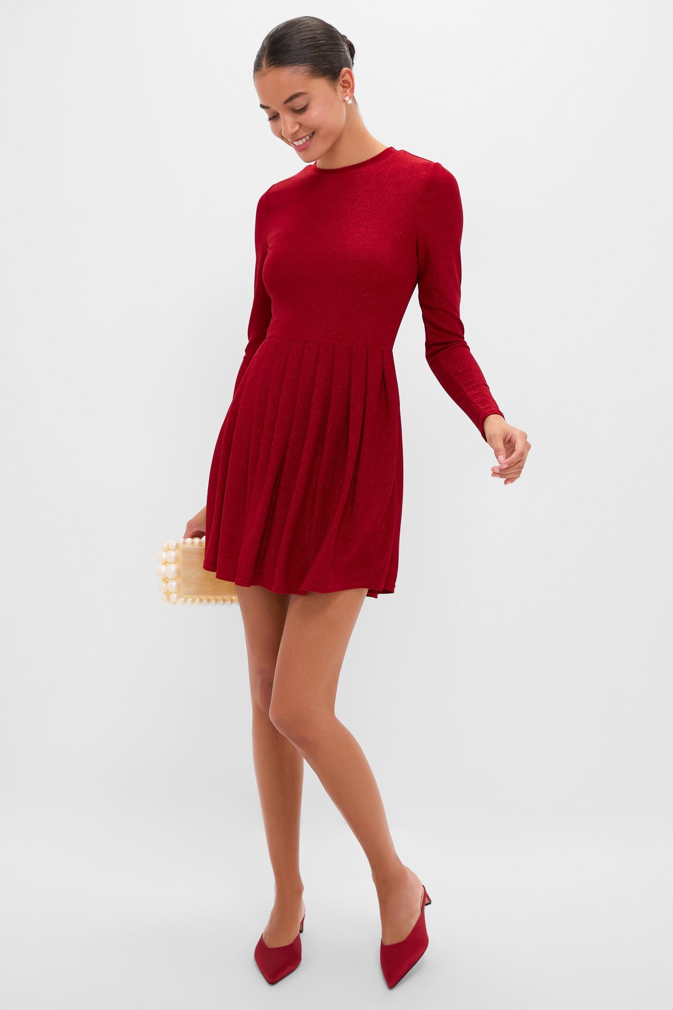 Woman in a classic Red Shimmer Pleated Bale Mini Dress, styled with an effortless bun, fun pearl-embellished clutch, and high-end red mules, exuding confidence.