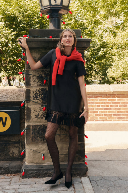 Woman wearing the classic Black Dupioni Tassel Downing Dress with fringe hem, sheer polka-dot tights, and heels, exuding effortless, fun elegance against a stone lamp post.
